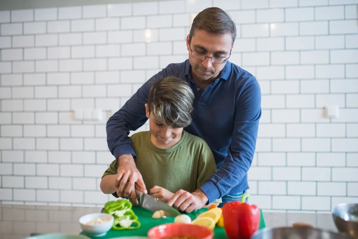 A father helps his son chop vegetables in a kitchen setting.