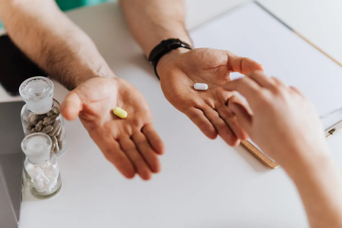 Hands showing two different pills with a background of medication bottles.