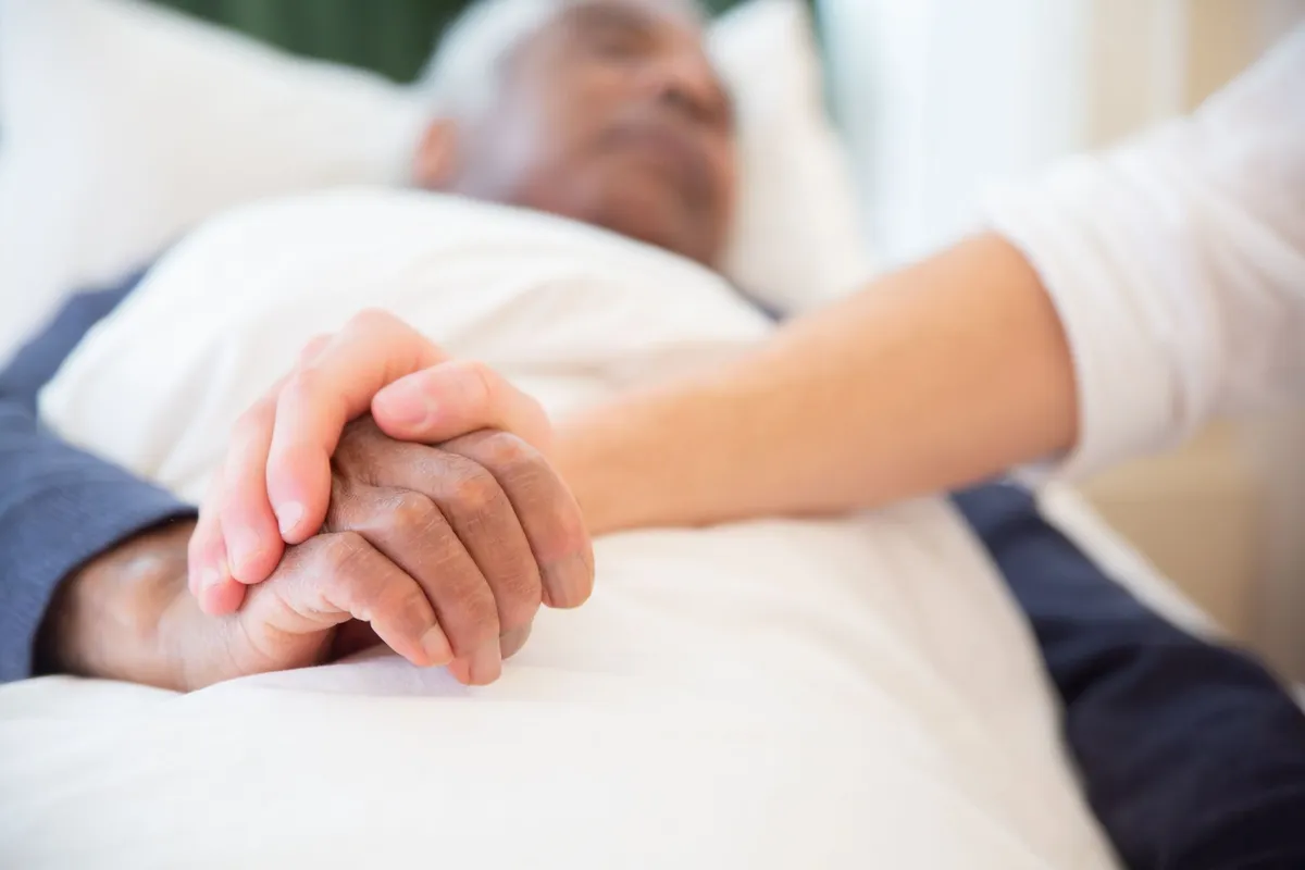 Two hands clasped together, one of an elderly person and one of a caregiver.