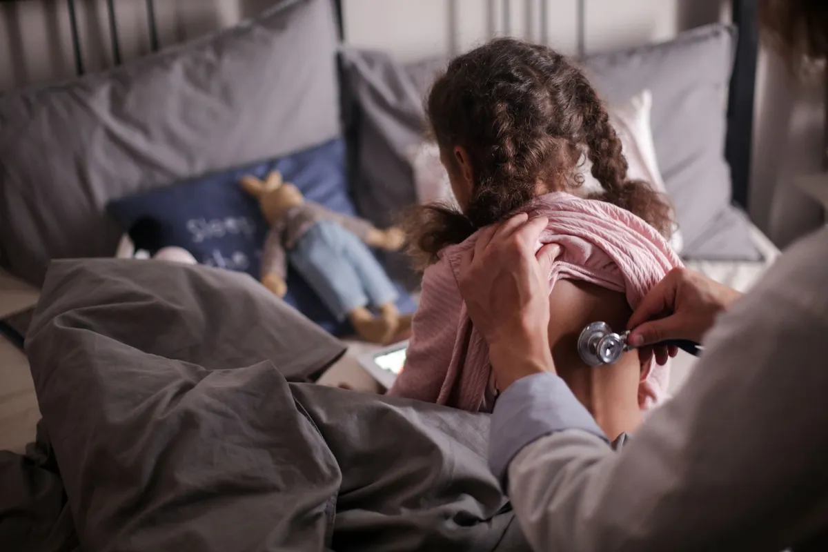 Doctor using a stethoscope on a child in bed with toys nearby.