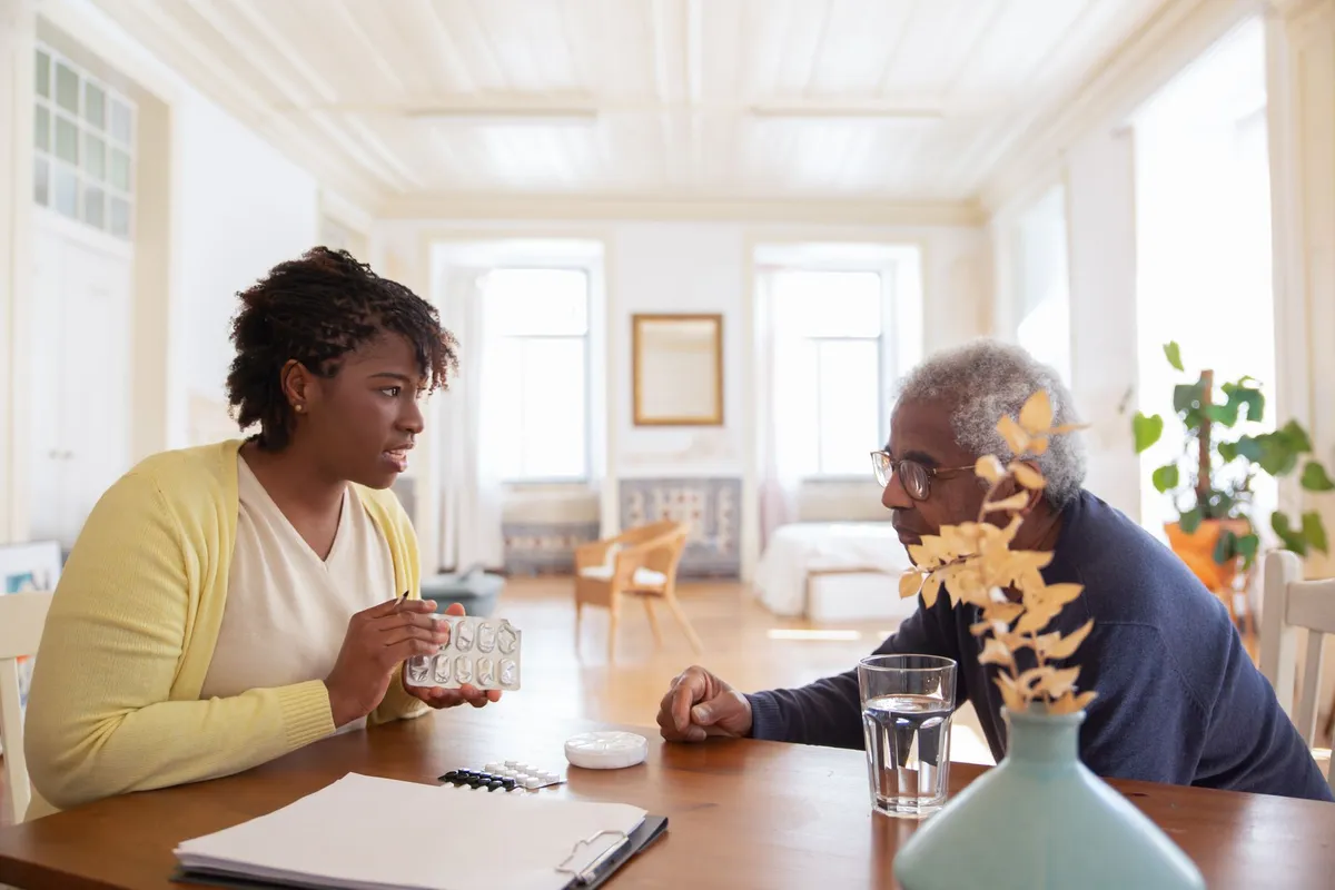 A caregiver discussing medication with an elderly man at a table in a bright room.