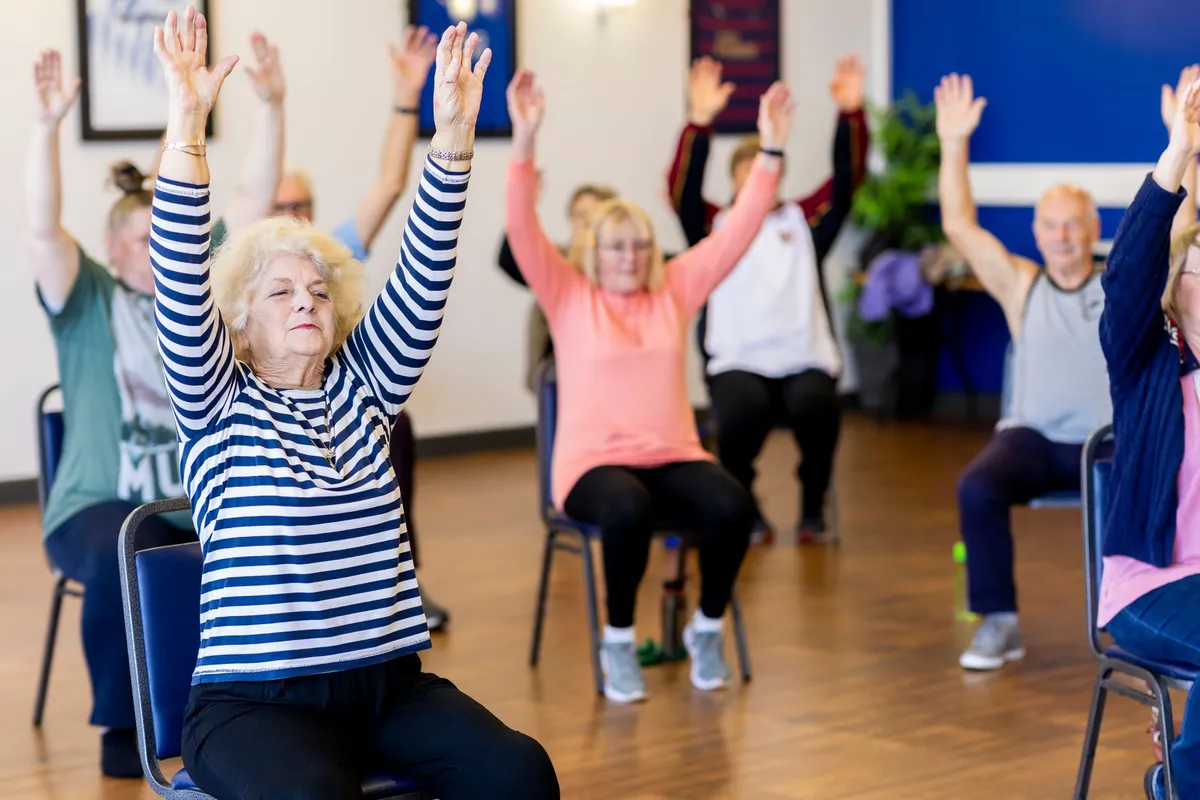 Seniors in a fitness class with arms raised, participating in seated exercises.