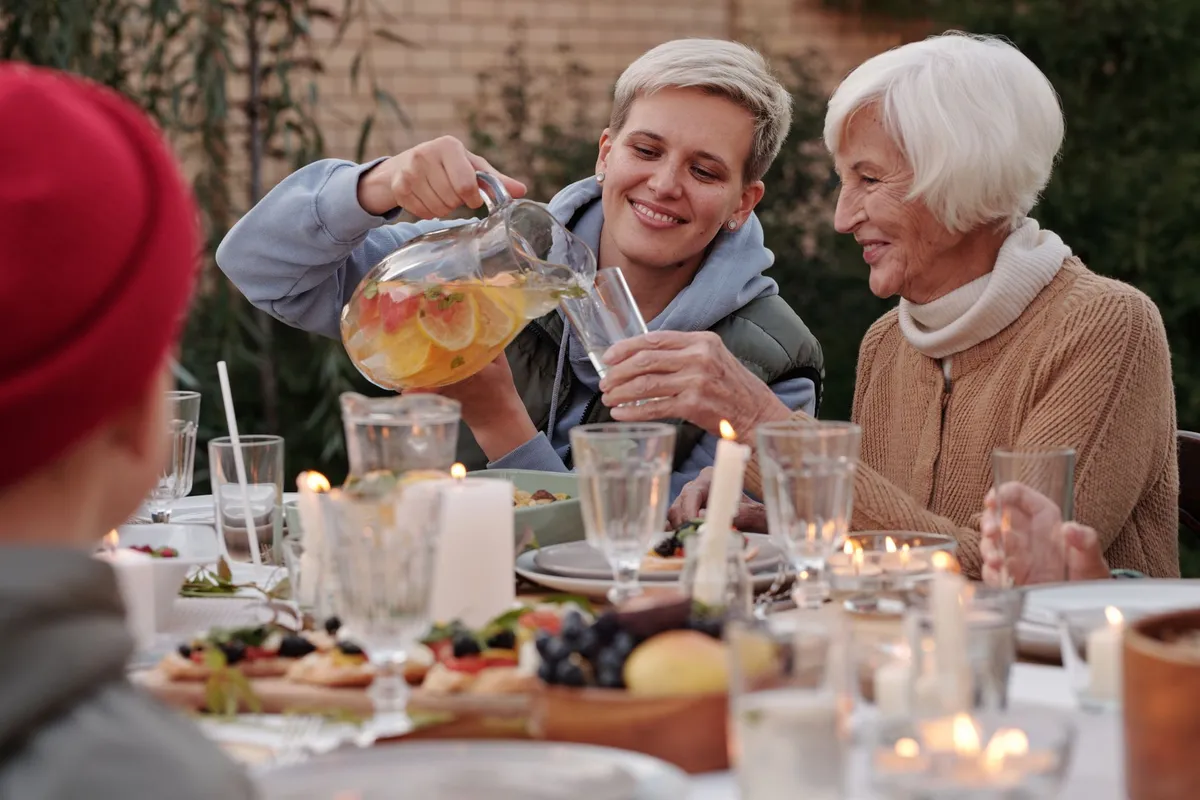 A young woman pours a drink for an elderly woman at a family meal with fruit and candles.