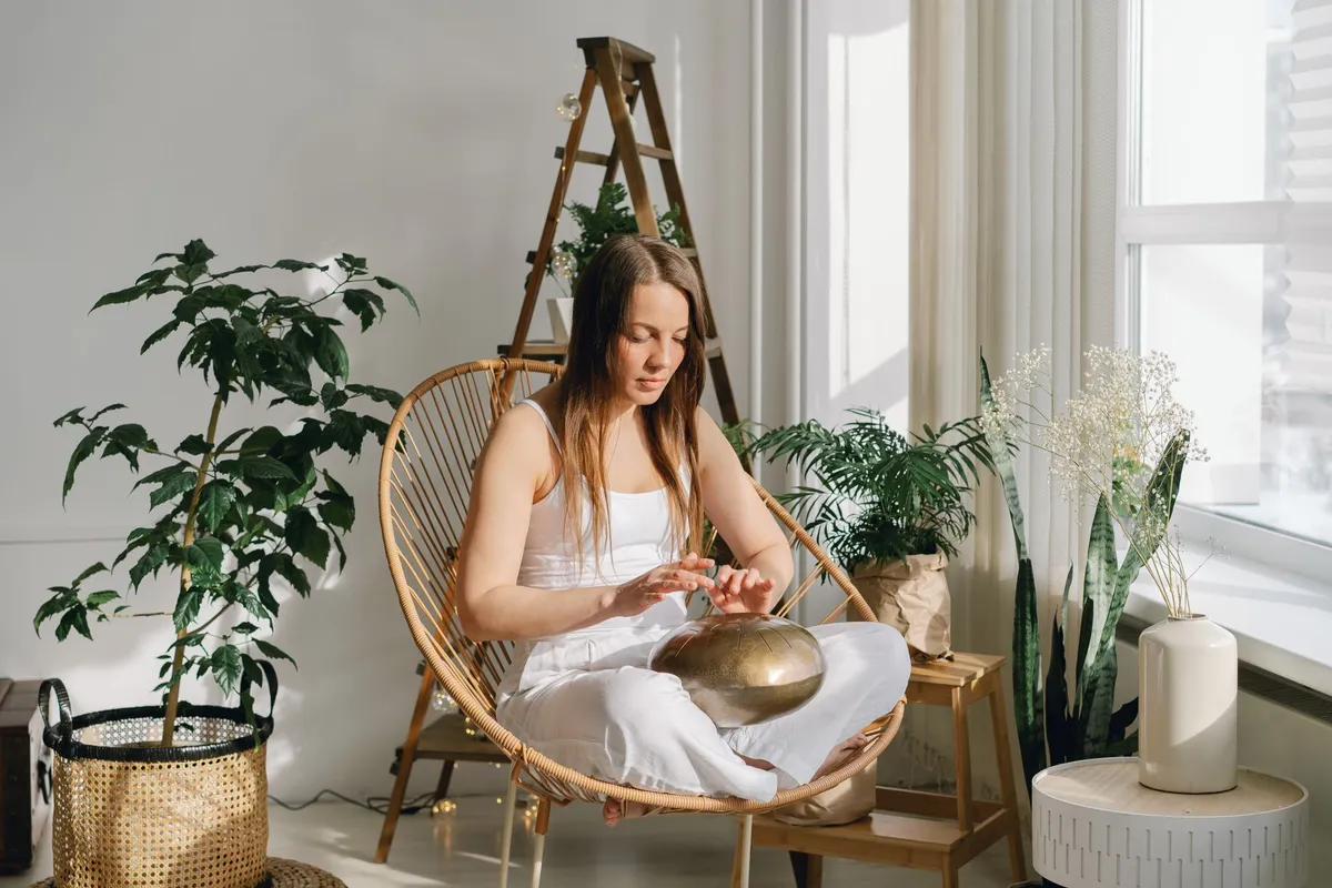 Person sitting in a chair, playing a singing bowl in a serene indoor space with plants.