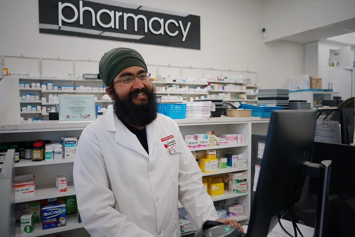 Pharmacist smiling at the counter in a pharmacy with medication shelves behind him.