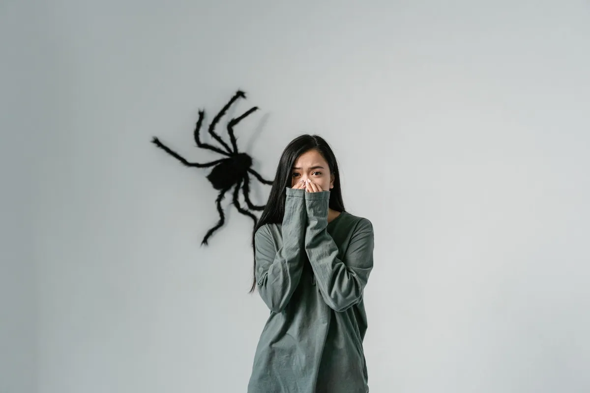 A girl covering her mouth in fear with a large spider silhouette on the wall behind her.
