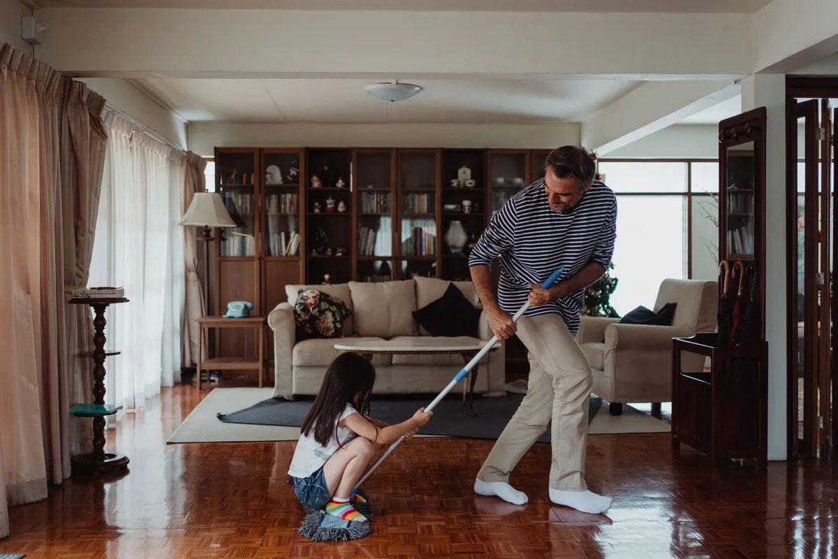 A man and a girl cleaning a living room with a mop and broom.