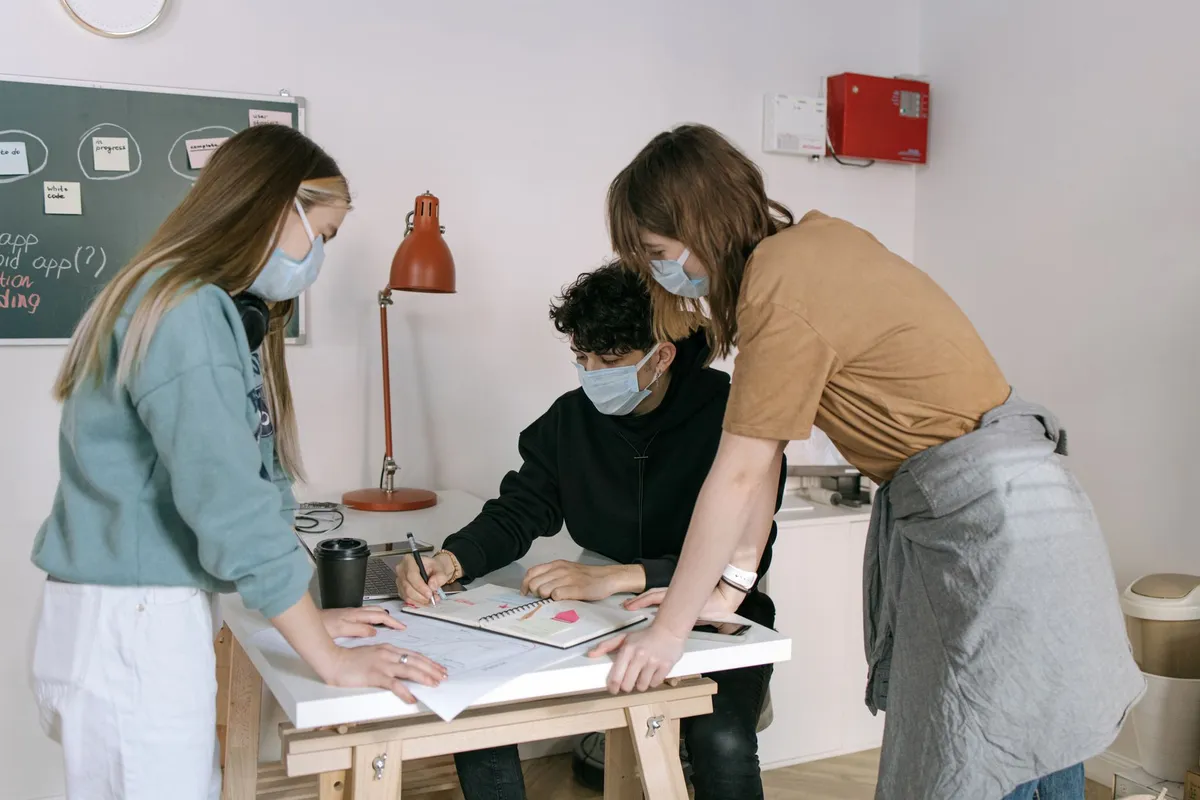 Three coworkers discussing plans at a table with notes and a laptop, wearing masks.