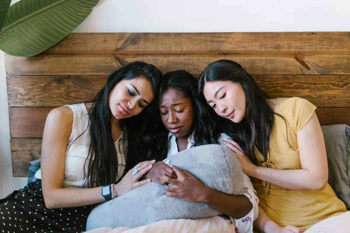 Three women comforting another woman who appears distressed, sitting on a bed.