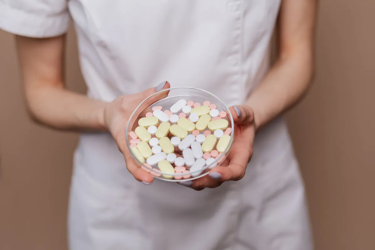 A person in a white coat holds a dish filled with various pills and capsules.