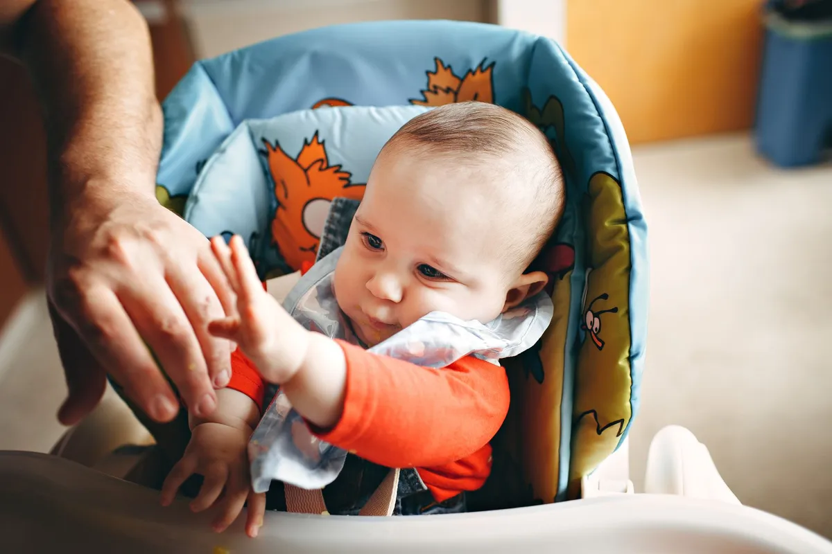 Baby in a highchair reaching for food with a caregiver’s hand nearby.