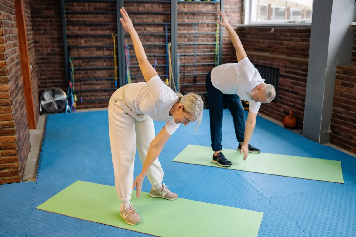 Two seniors stretching to the side on exercise mats in a fitness setting.