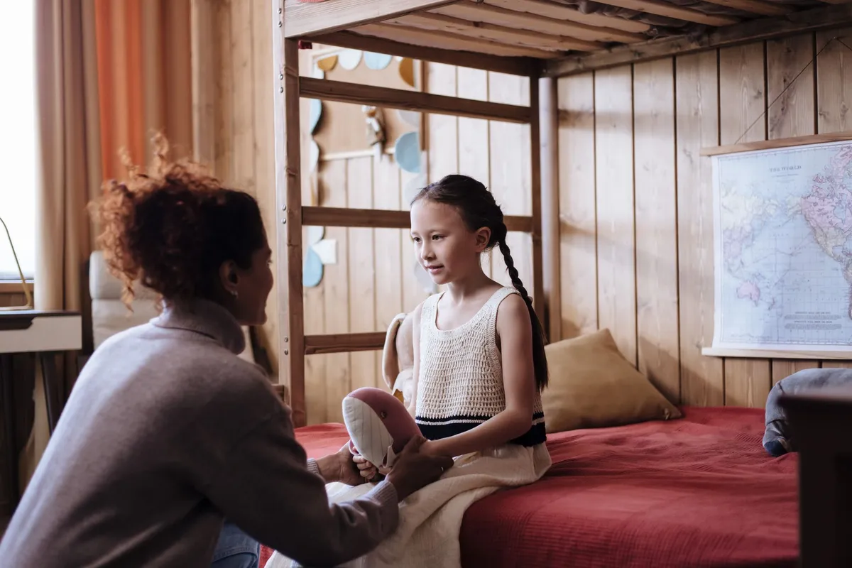 A caregiver comforting a child in a bedroom setting, discussing feelings after bedwetting.