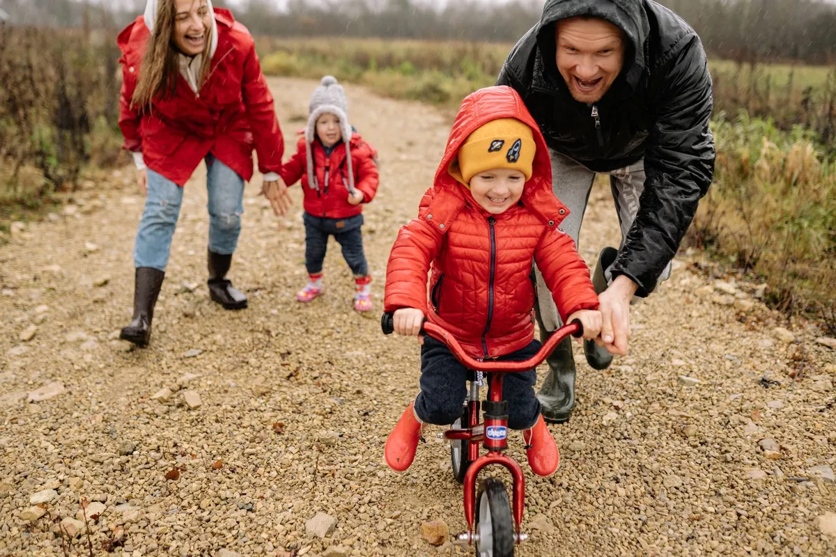 Family encouraging a child riding a bike on a gravel path in a rural setting.