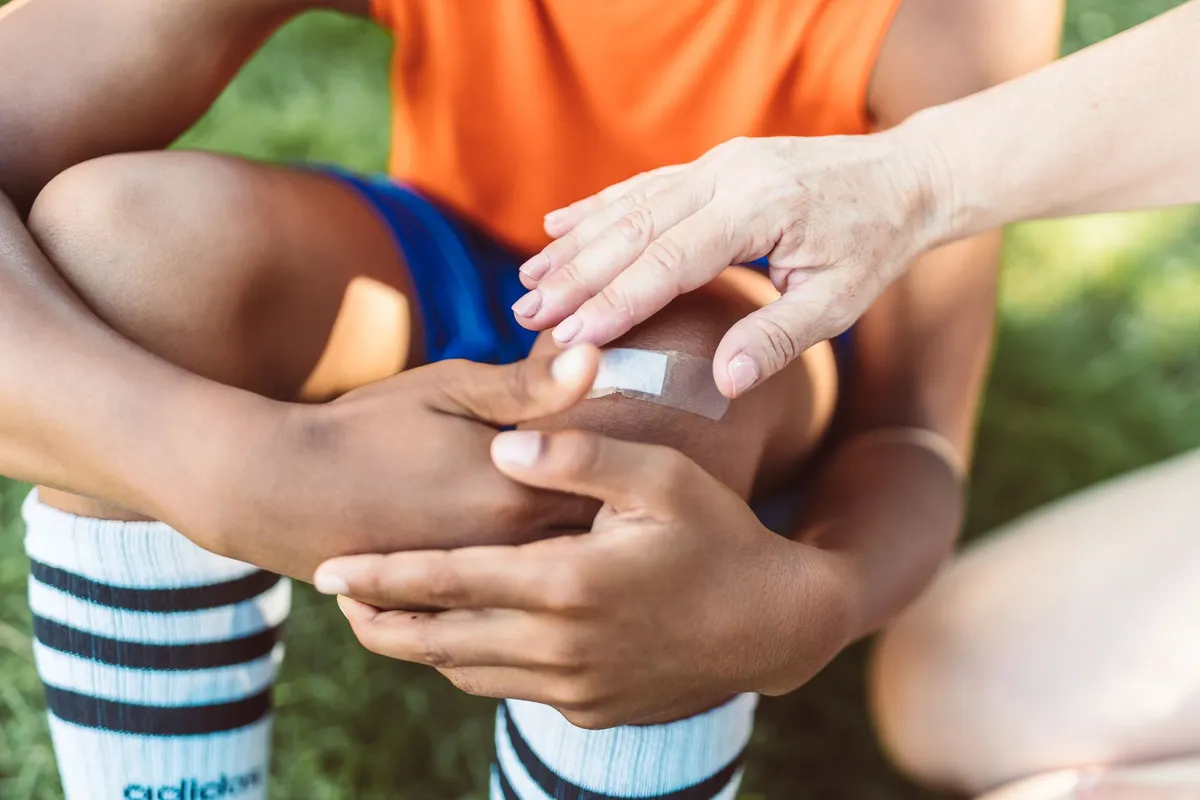 A person applying a bandage to a child’s knee after an injury in a grassy area.
