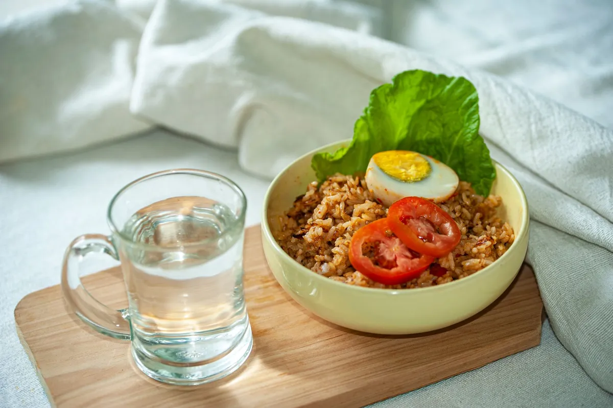 A bowl of rice with vegetables and a glass of water on a wooden board.