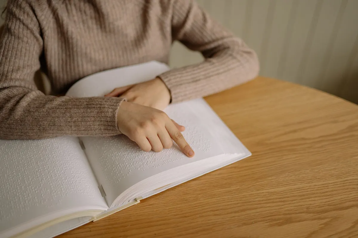 Hand pointing at a page of braille book on a wooden table.