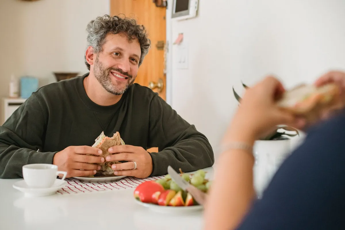 Man smiling while holding a sandwich at a breakfast table with fruit and coffee.