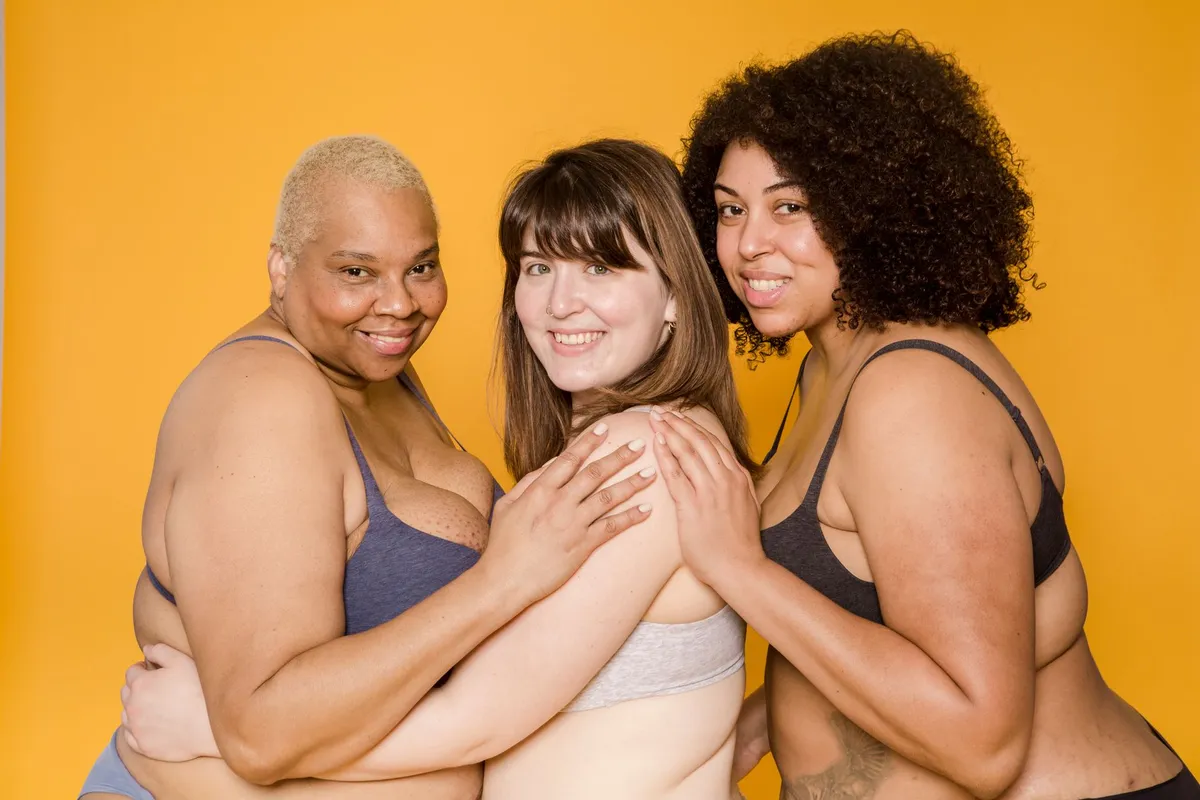 Three women in bras posing together against a yellow background.