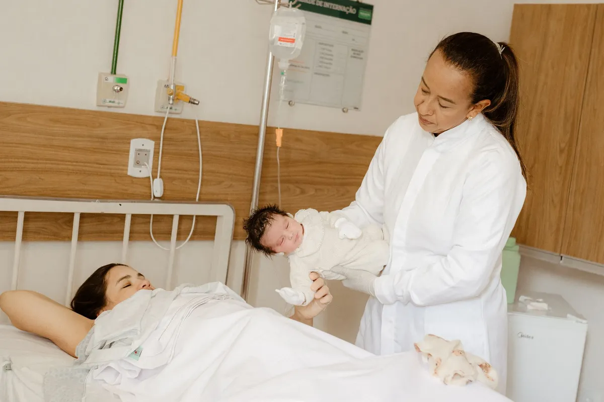 A nurse holds a newborn baby while a mother rests in a hospital bed.