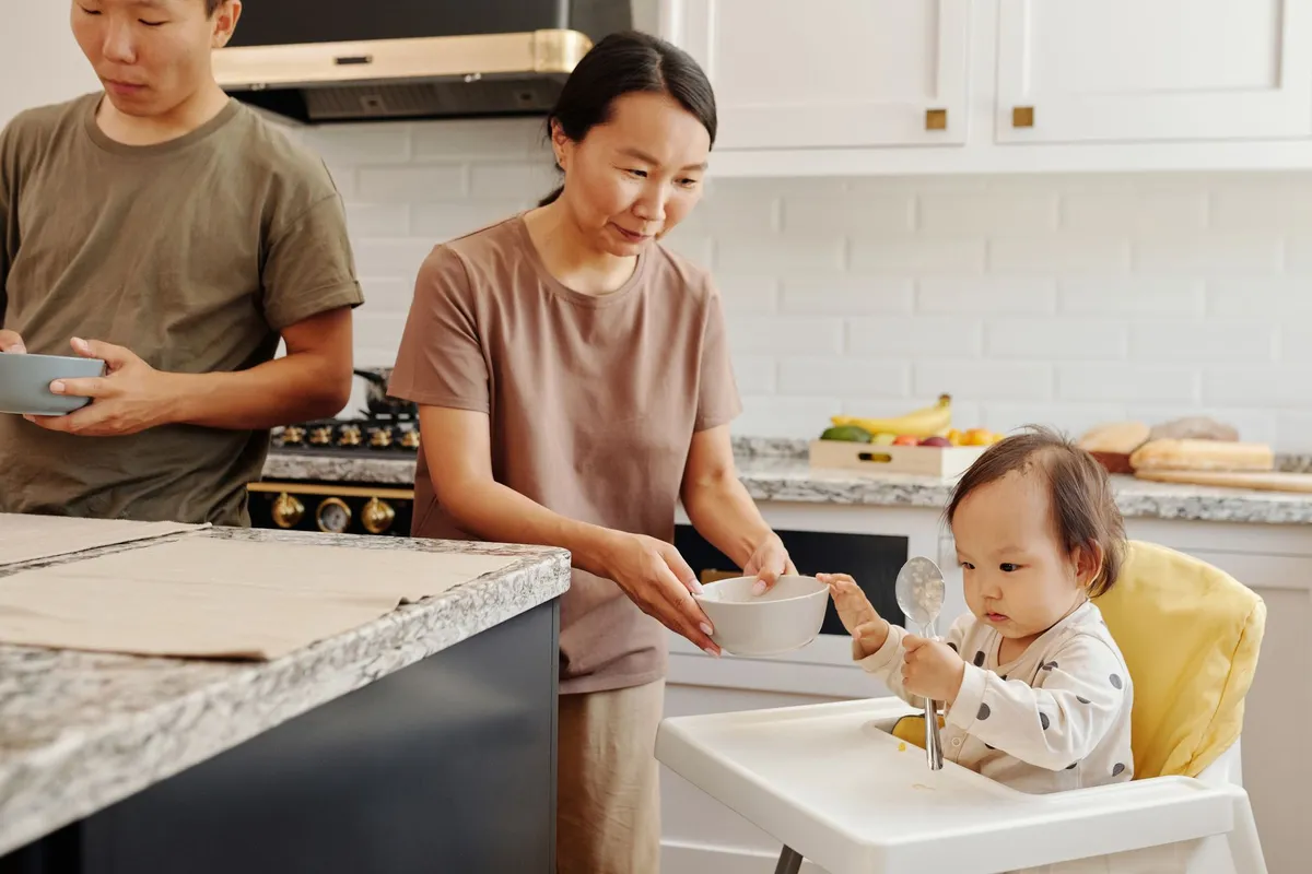 Parent serving food to a child in a high chair in a kitchen setting.