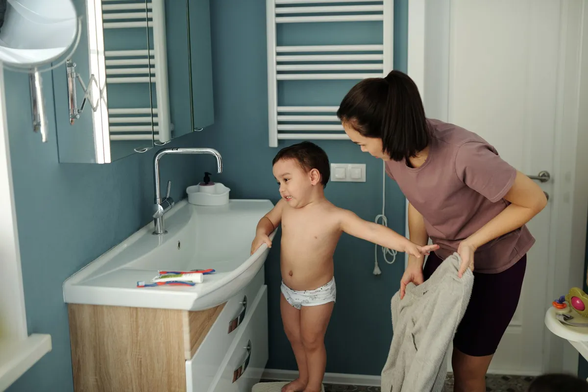 Child in a bathroom with a parent nearby, preparing for a bath.