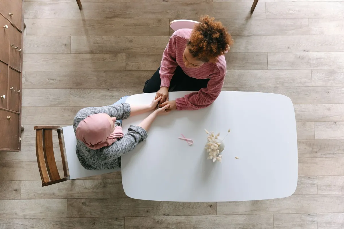 Two women holding hands at a table, one wearing a headscarf, symbolizing support during cancer treatment.