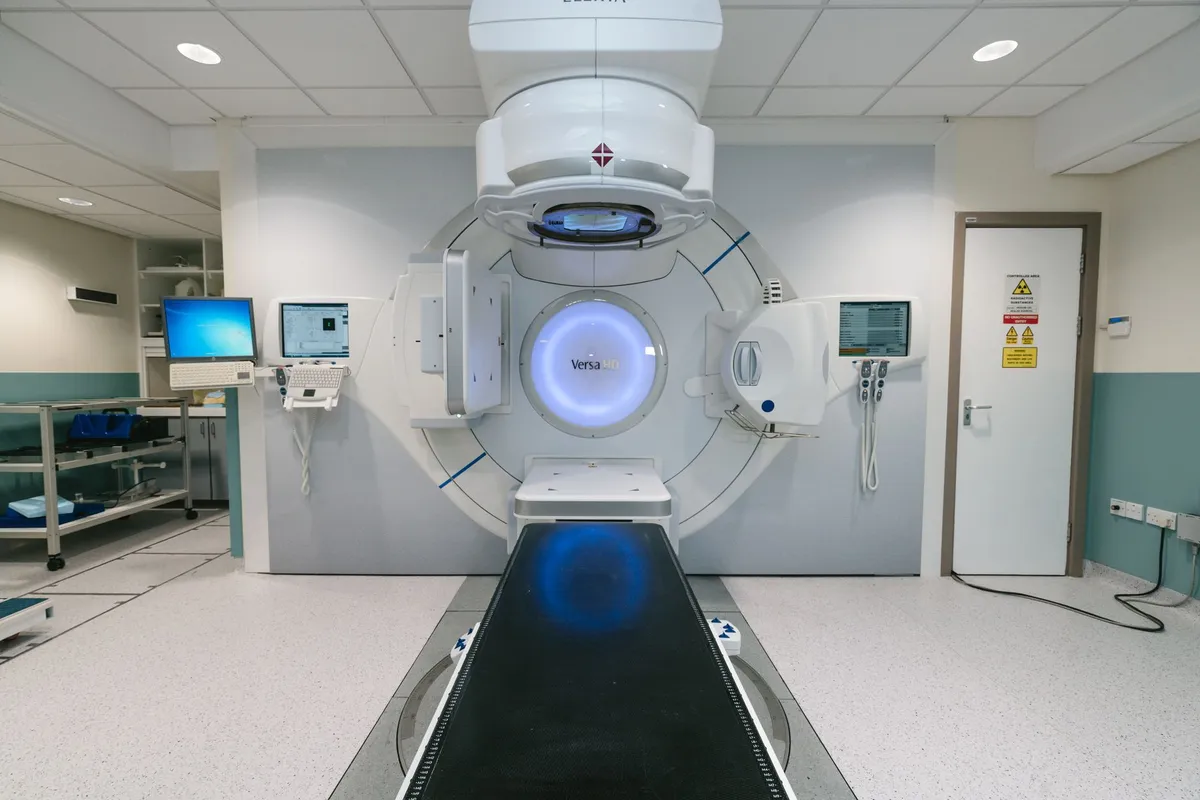 Radiotherapy machine in a clinical setting with a treatment table and monitors.