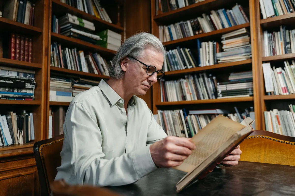 Man reading an old book in a library with shelves of books behind him.