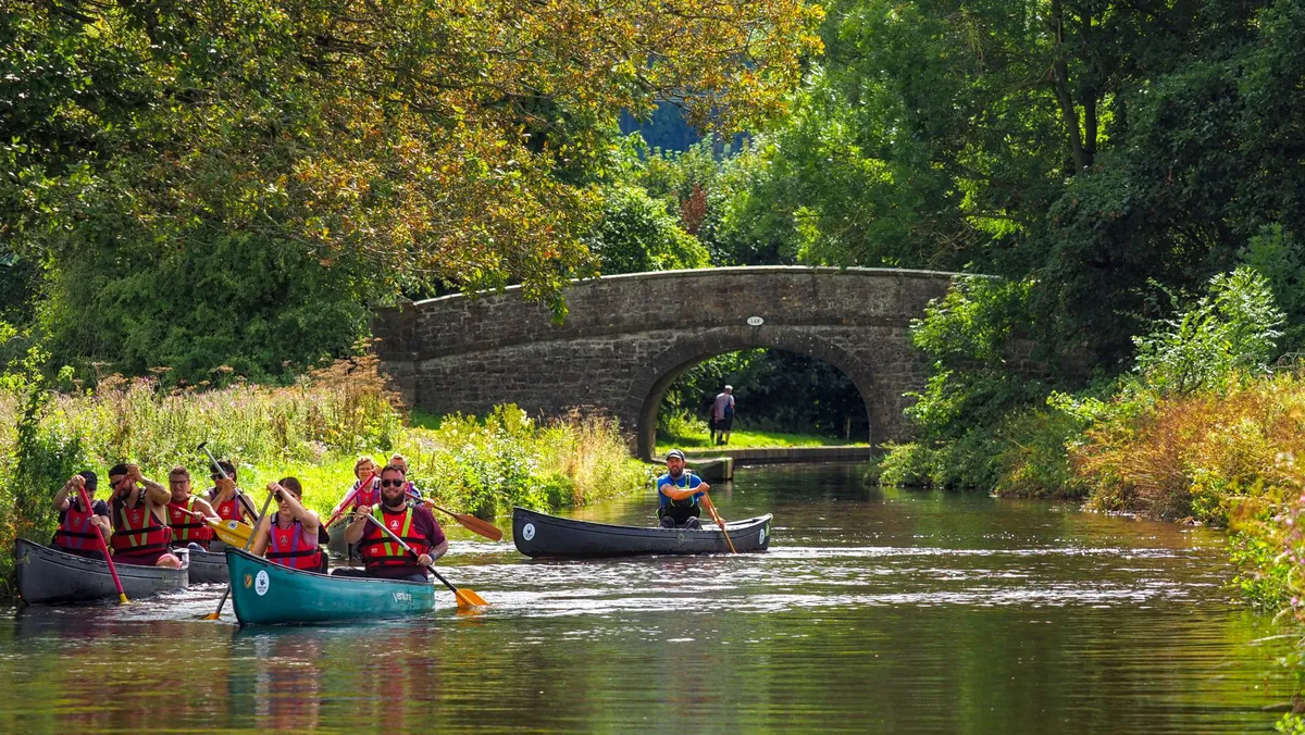 Group of people canoeing on a calm waterway with greenery and a bridge in the background.