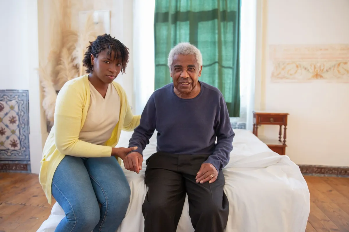 Caregiver assisting elderly man sitting on a bed, showing support and companionship.