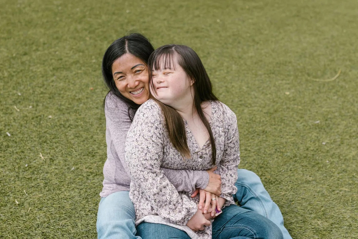 Two women sitting on grass, one embracing the other with smiles.