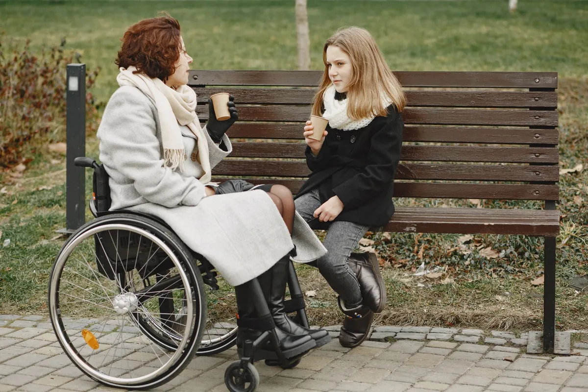 Woman in wheelchair talking with a girl on a park bench, both holding cups.