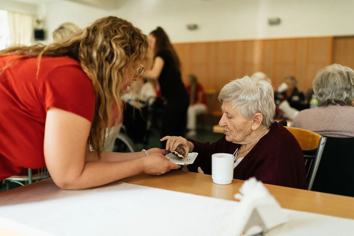 Caregiver assisting elderly woman at a table in a communal setting.