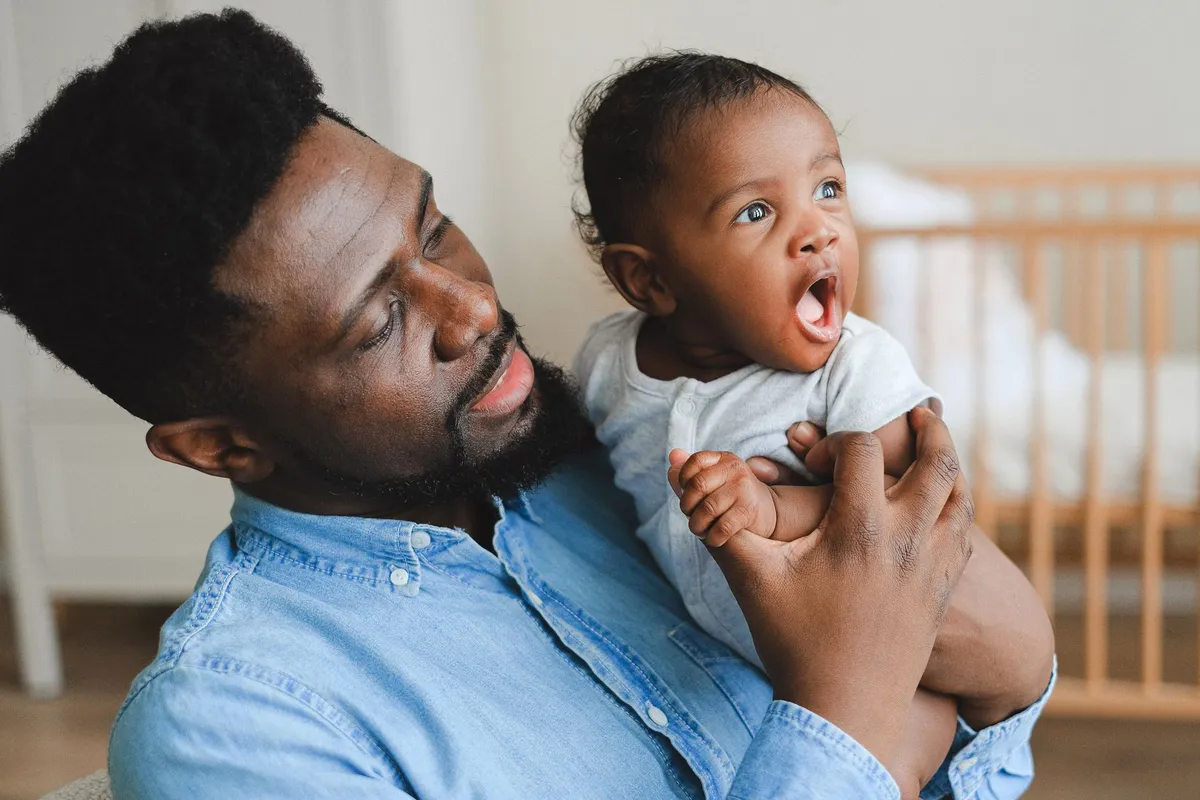 A father holding and smiling at his yawning six-month-old baby indoors.