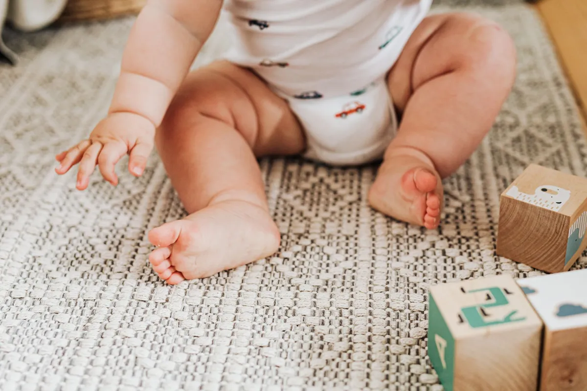 Baby sitting on a rug reaching for wooden blocks with bare feet visible.