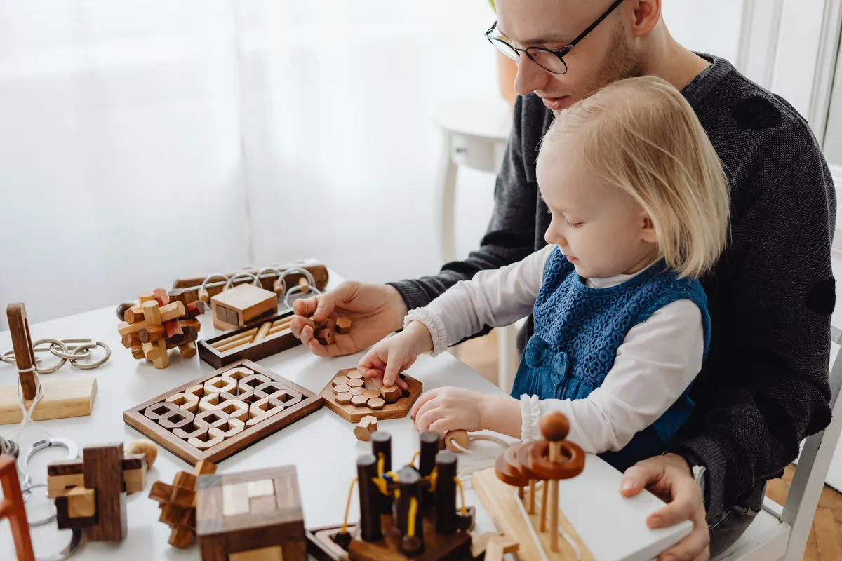 Toddler playing with wooden toys alongside an adult in a bright room.