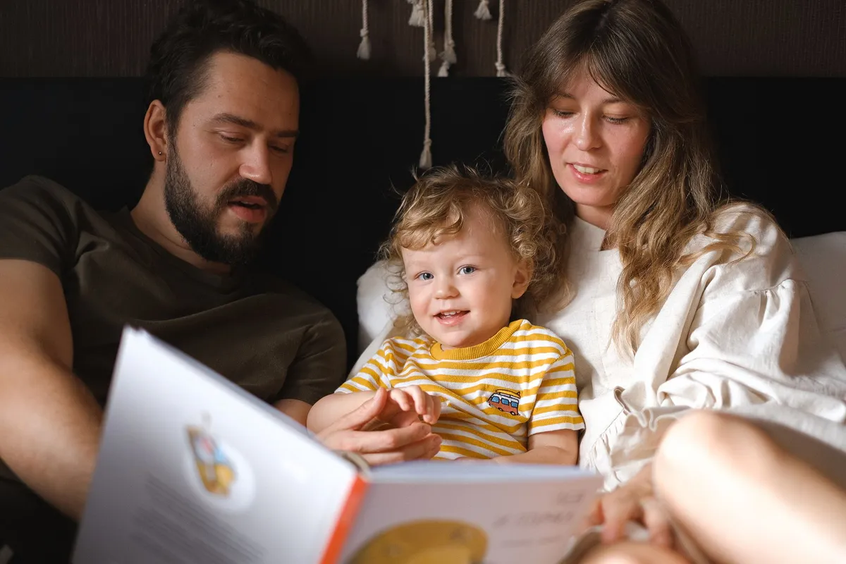 Parents reading a book with a smiling young child in bed.
