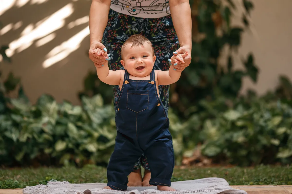 Toddler standing with assistance, smiling, in a garden setting.