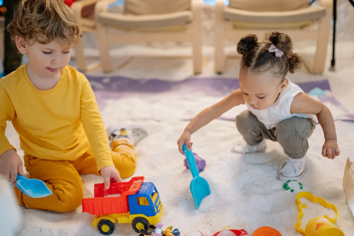 Two toddlers playing with toy trucks and shovels in a sand-like environment.