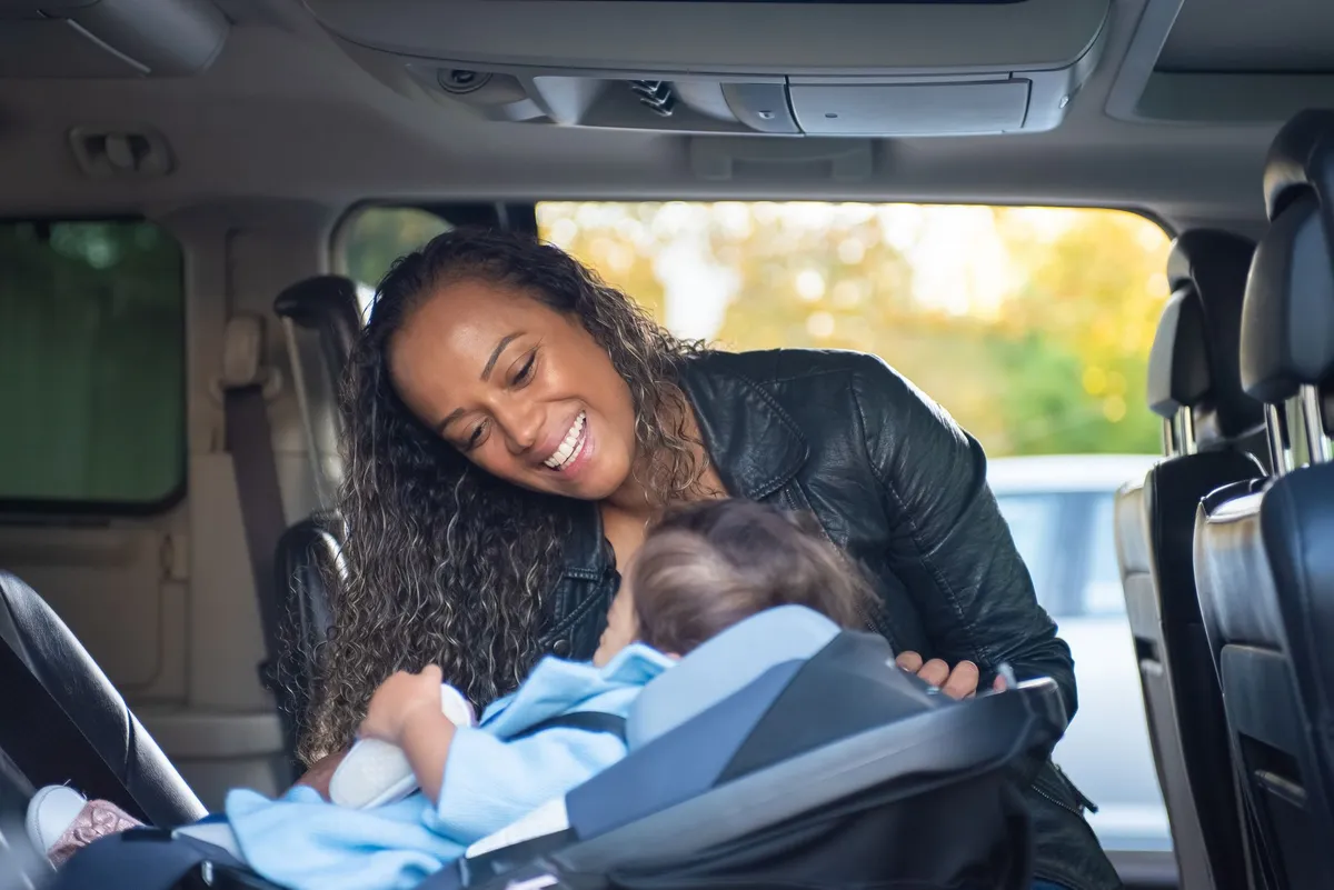 A smiling woman interacts with a baby in a car seat inside a vehicle.