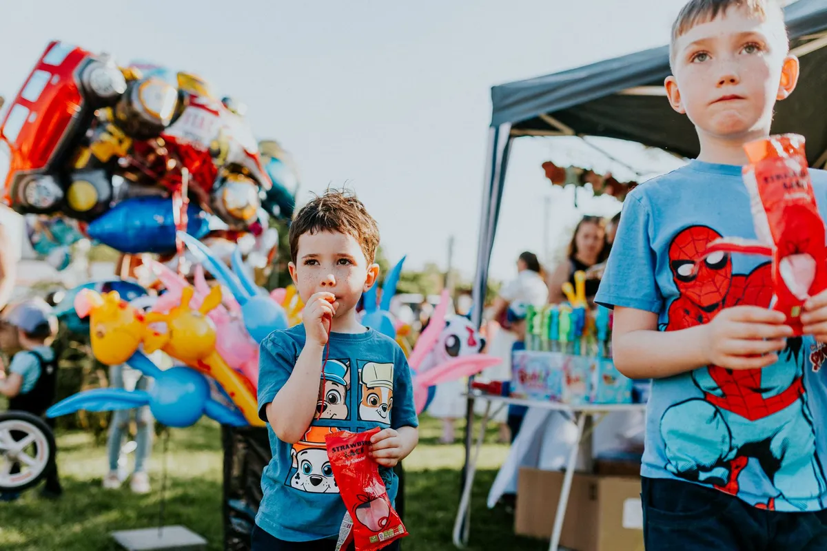Two children enjoying snacks at an outdoor event with colorful balloons in the background.