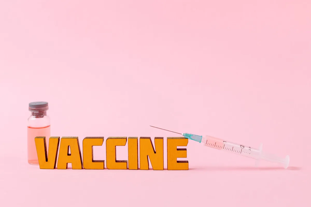 Colorful letters spelling ‘VACCINE’ next to a syringe and vaccine vial on a pink background.