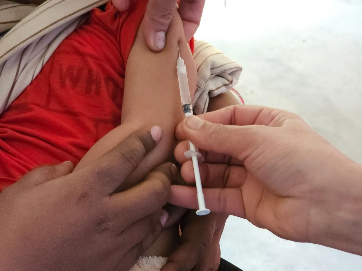 Child receiving a vaccination in a clinic setting with a caregiver’s hand visible.
