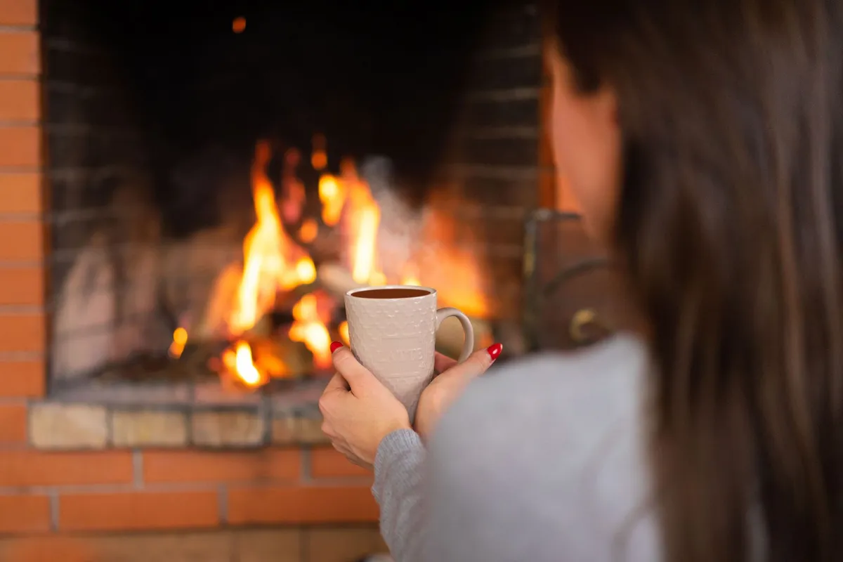 Woman holding a mug in front of a fireplace, suggesting warmth during cold weather.