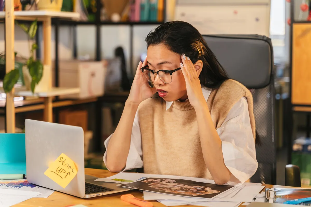 Stressed employee at desk with hands on head, surrounded by papers and a laptop.