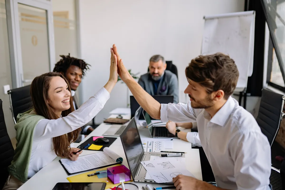 Two colleagues high-fiving in a collaborative office setting with others working nearby.