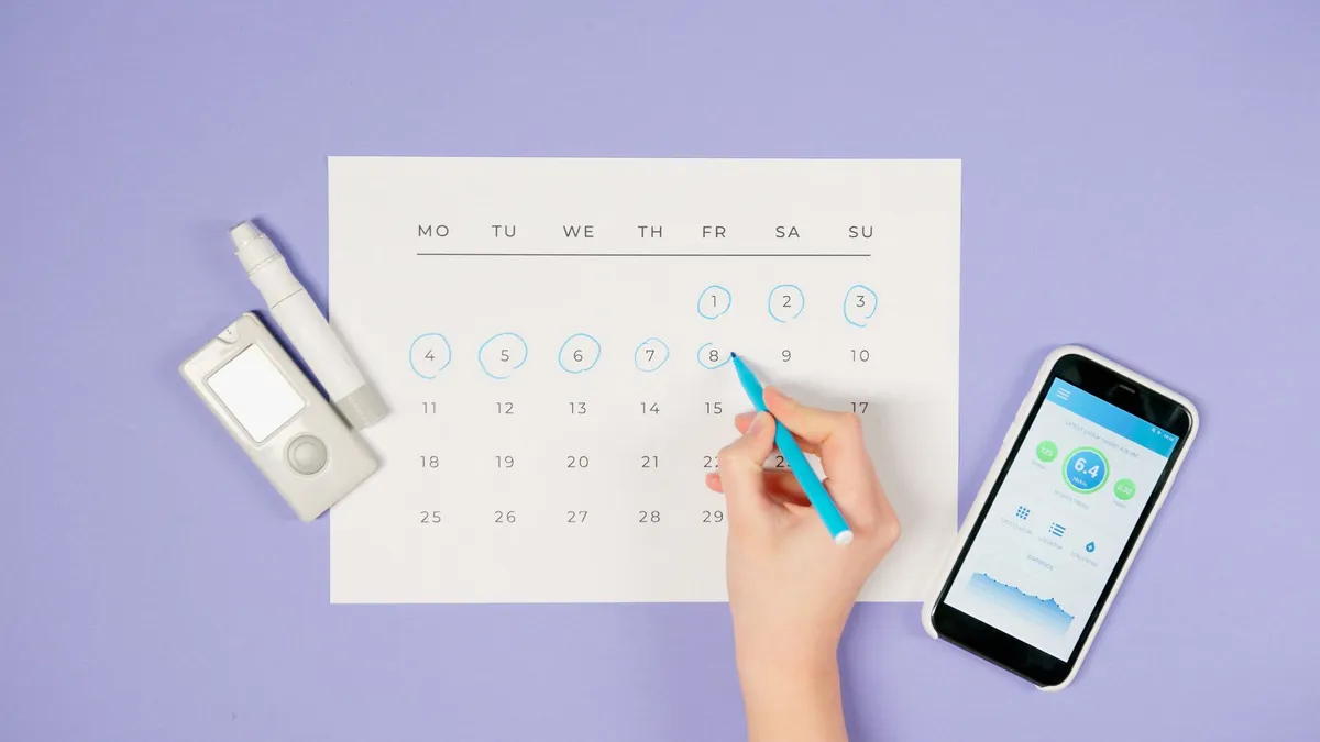 Hand marking dates on a calendar with a phone and ovulation tracker nearby.