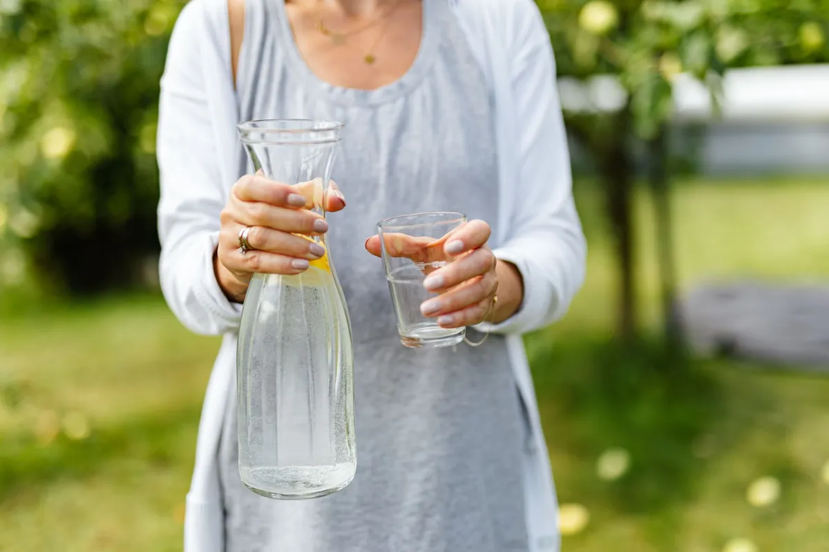 Person holding a pitcher of water and a glass in a garden setting.