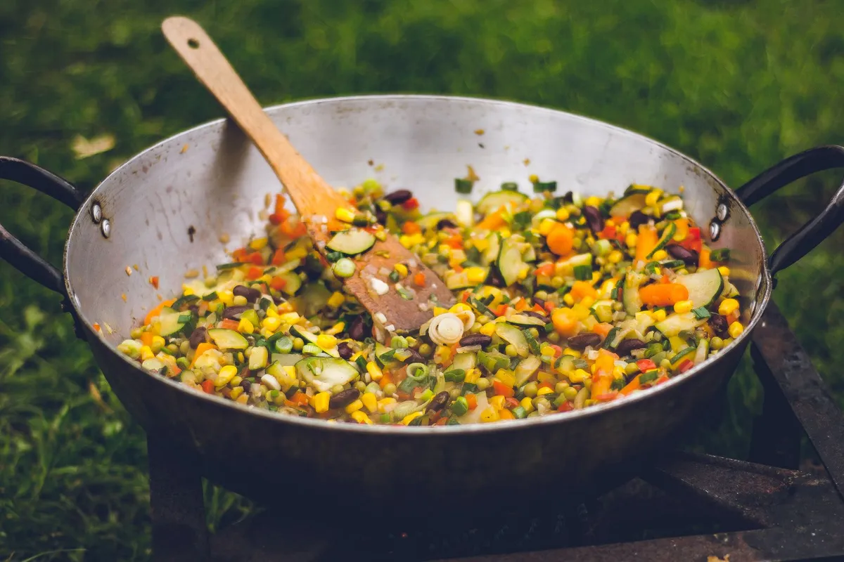 Colorful vegetable stir-fry in a large pan with a wooden spoon outdoors.